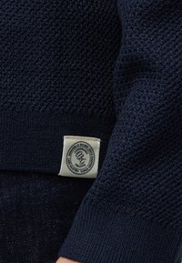 Manche de pull en tricot bleu foncé avec une étiquette en tissu blanche affichant un texte circulaire et un logo près du poignet.
