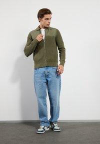 Maslinasta zip-up cardigan s rebrastom teksturom, kontrastnim ramenim panelima, uparen s širokim plavim trapericama i tamnim tenisicama.