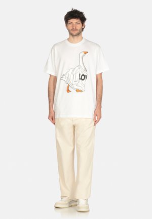 Uomo che indossa una maglietta bianca con una grafica di un'oca avvolta in un panno, pantaloni beige e sneaker beige, in piedi su uno sfondo neutro.