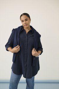 Femeie purtând o cămașă bleumarin cu nasturi și un pulover asortat așezat pe umeri, stând în fața unui fundal simplu și deschis la culoare.