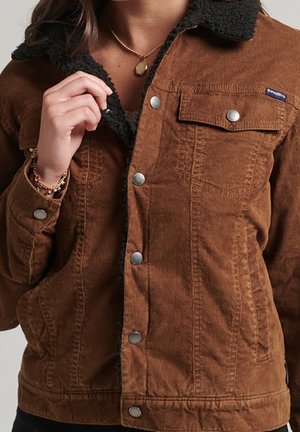 Chaqueta de pana marrón con cuello de forro polar negro, botones de presión plateados, bolsillo en el pecho y un collar con colgante dorado en la persona que la lleva puesta.