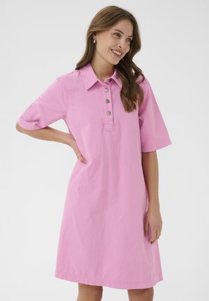 Vrouw die glimlacht met hand op heup, gekleed in een roze kantoorkleed met korte mouwen en een kraag, tegen een effen lichte achtergrond.