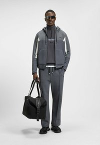 Ensemble de sport gris composé d'un sweat à capuche zippé et de pantalons ajustés, fabriqué en tissu texturé. Sac de sport noir avec fermeture éclair, porté à l'épaule.