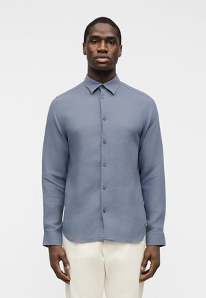 Άνδρας στέκεται προς τα εμπρός φορώντας μπλε πουκάμισο με κουμπιά και μακριά μανίκια και ανοιχτόχρωμο παντελόνι με φόντο μία απλή λευκή επιφάνεια.