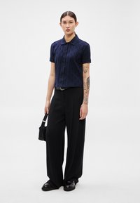 Tumši zila īsroku krekls ar teksturētu rakstu, kopā ar augstu jostasvietu melniem plaša piegriezuma biksēm un melniem apaviem. Mazs melns somiņa.