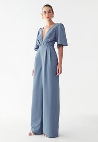 Modra kombinezona z globokim V-odskim in kratkimi naborki. Izdelana iz strukturirane tkanine, z širokimi hlačnicami in prilegajočim pasom. Minimalističen dizajn.