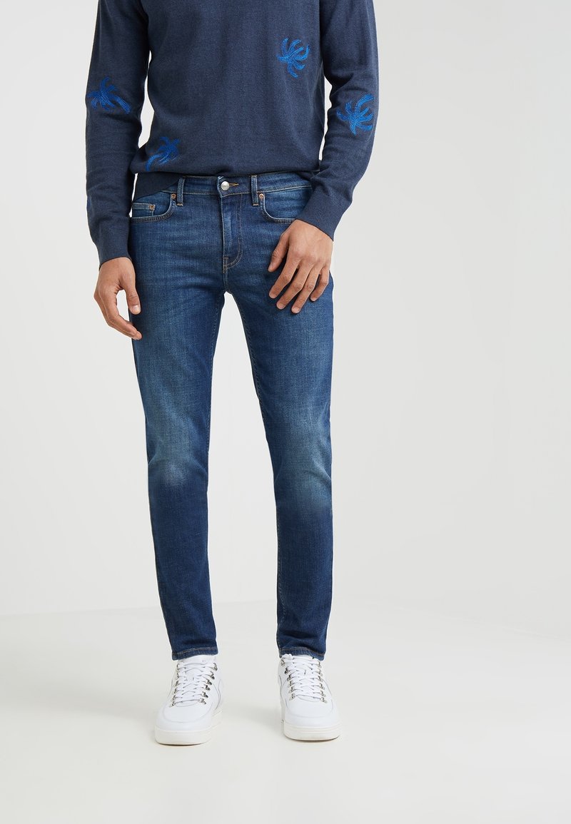 Homme portant un jean slim bleu en denim, des baskets blanches et un pull bleu marine avec des motifs brodés bleus sur fond blanc.