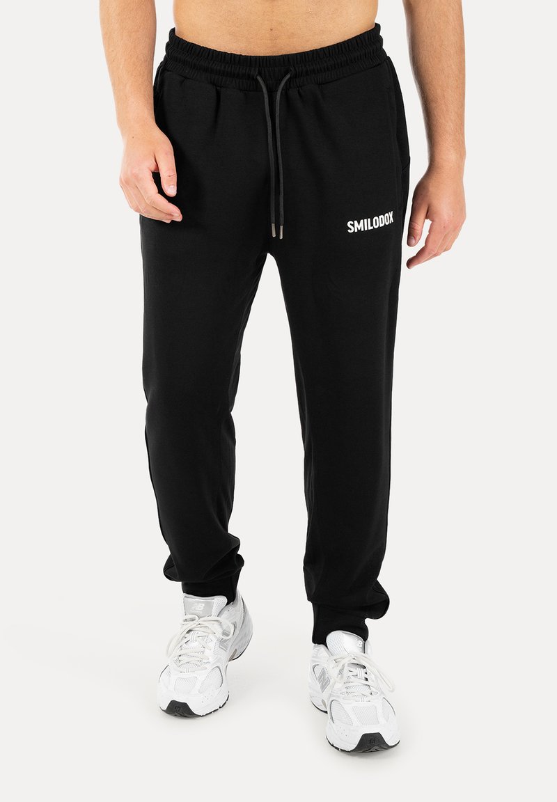 Homme portant un pantalon de jogging noir avec cordon et logo "SMILODOX", associé à des baskets blanches sur fond blanc.