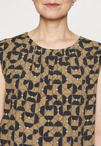 Bruine blouse met een geprint patroon, bestaande uit een geometrisch ontwerp met zwarte en zachte groene accenten. De blouse heeft een ronde halslijn en geplooide details aan de voorkant.