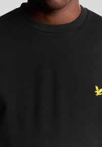 T-shirt nero a maniche corte con scollatura rotonda, caratterizzato da un piccolo logo di un uccellino giallo sulla parte sinistra del petto. Texture liscia e uniforme.