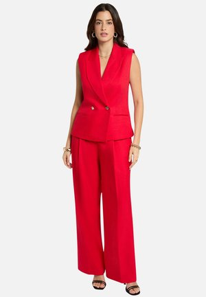 Kvinde iført rød ærmeløs blazer med dobbelte knapper og matchende bukser med vide ben, accessorized med guldsmykker og sorte sandaler.