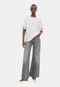 T-shirt bianca in cotone con dettagli in pizzo sulle maniche, abbinata a jeans grigi a gamba larga e scarpe bianche a punta. Design semplice e moderno.