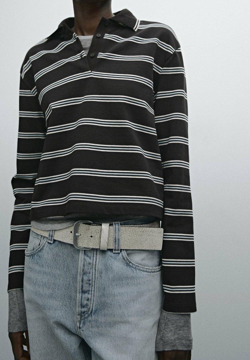 Personne portant un t-shirt à manches longues rayé noir et blanc par-dessus des manches grises, un jean bleu clair, et une ceinture gris clair texturée avec une boucle métallique.