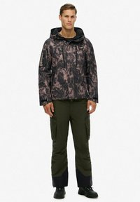 Man staat gekleed in een zwart en bruin gestreept hoodiejack, olijfgroene cargo-broeken met zwarte onderstukken, en zwarte schoenen op een witte achtergrond.