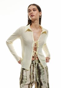 Cardigan giallo chiaro e trasparente con colletto. Presenta maniche lunghe, scollatura a V profonda e chiusura anteriore con fiocco. Tessuto testurizzato con motivi sottili.