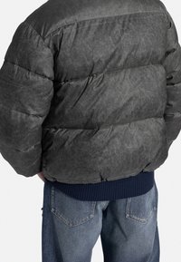 Persona che indossa una giacca a vento grigia scura e jeans blu, mostrata di spalle su uno sfondo bianco uniforme.