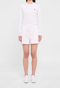 Chemise blanche à manches longues avec un petit logo bleu marine, associée à un short beige clair et des baskets blanches, posant devant un fond uni.