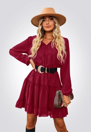 Vestido burdeos de manga larga con gotas texturizadas, dobladillo con volantes y un cinturón negro con hebilla plateada. Bolso marrón con cadena.