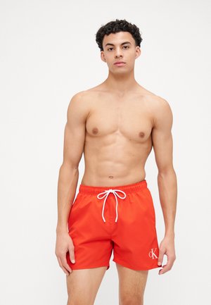 Red swim trunks made of lightweight material with an elastic waistband and white drawstring, featuring a small "CK" logo on the side.