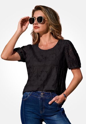Frau trägt schwarze kurzärmlige Bluse mit floralem Muster, blaue Jeans, Sonnenbrille und Armband, posiert mit einer Hand in der Tasche und einer Hand, die die Brille anpasst.