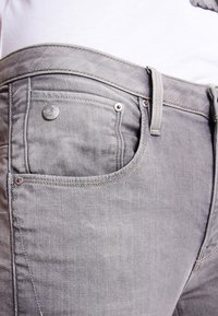 Grå jeans med en slät yta, med en framficka, metallknapp och kontrasterande sömmar. Midjebandet är något vikt.