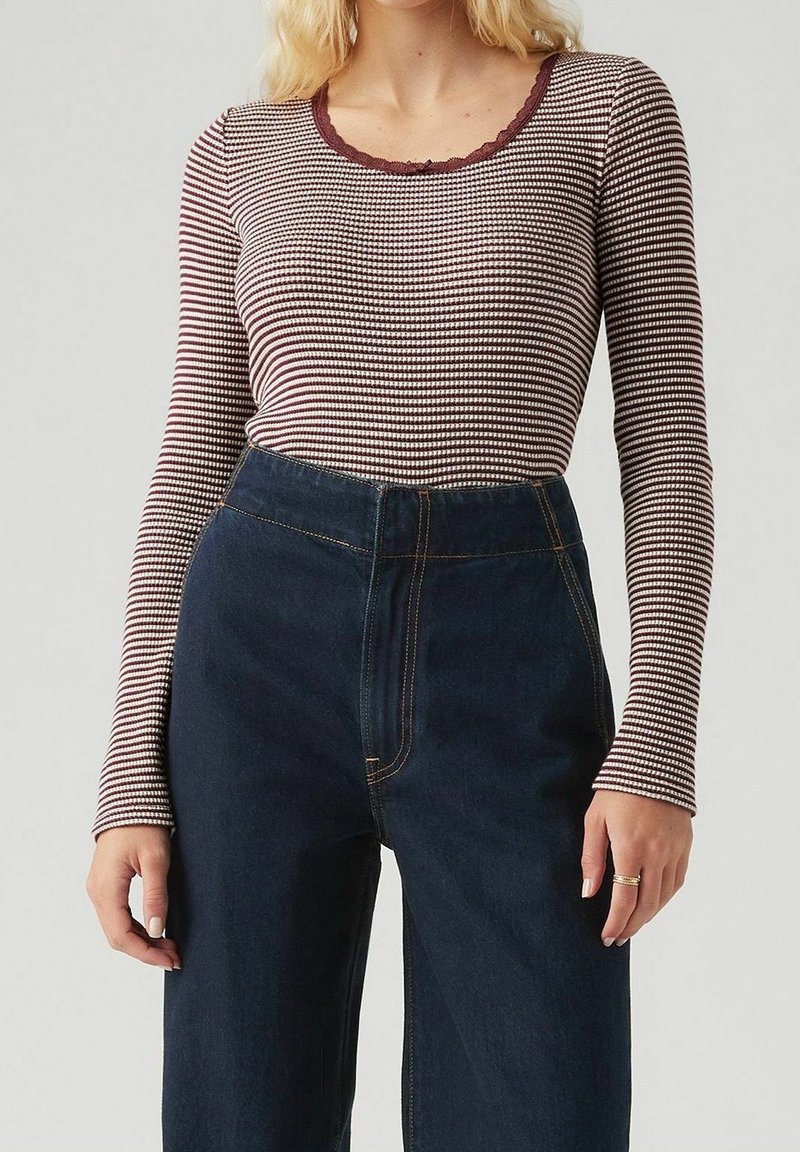 Femme portant un haut à manches longues rayé bordeaux et blanc, rentré dans un jean taille haute bleu foncé avec des coutures contrastées.