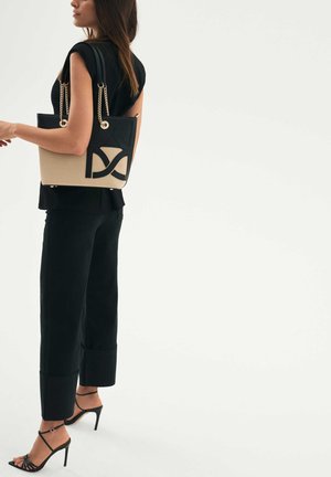 Mustan ja beige-kylkinen laukku, jossa on kaksinkertaiset yläkahvat, kultaketjuviimeistely ja geometristen leikkauskuvioiden muotoilu sivussa. Sulava materiaalipinta.