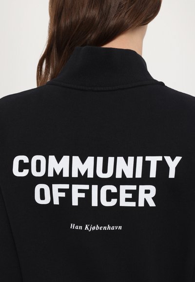Melna sporta krekls ar augstu apkakli. Uz priekšpuses ir izteikta balta teksta frāze "KOPIENS OFICIERIS", zem tās mazākā fontā "Han Kjøbenhavn". Mīksts auduma tekstūra.