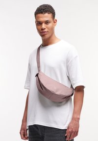 Borsa a tracolla in morbido tessuto rosa chiaro con forma curva e tracolla regolabile, indossata sopra una camicia bianca. Marchio discreto sul davanti.