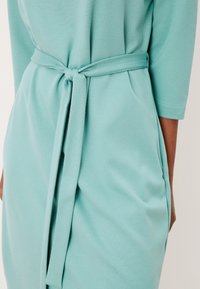 Abito verde menta con una cintura legata in vita, dotato di maniche a trequarti e di un tessuto morbido e texturizzato. Caduta morbida su entrambi i lati.