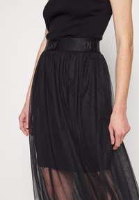 Musta tüllihame, jossa on istuva vyötärö ja "DKNY"-logo. Läpinäkyvä rakenne ja kerroksellinen muotoilu luovat virtaavan siluetin.