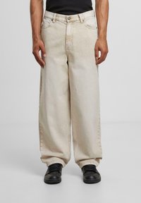Beige wide-leg jeans made of denim with a faded wash, featuring five pockets and silver hardware. Paired with black sneakers.