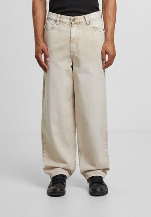 Beige wide-leg jeans made of denim with a faded wash, featuring five pockets and silver hardware. Paired with black sneakers.