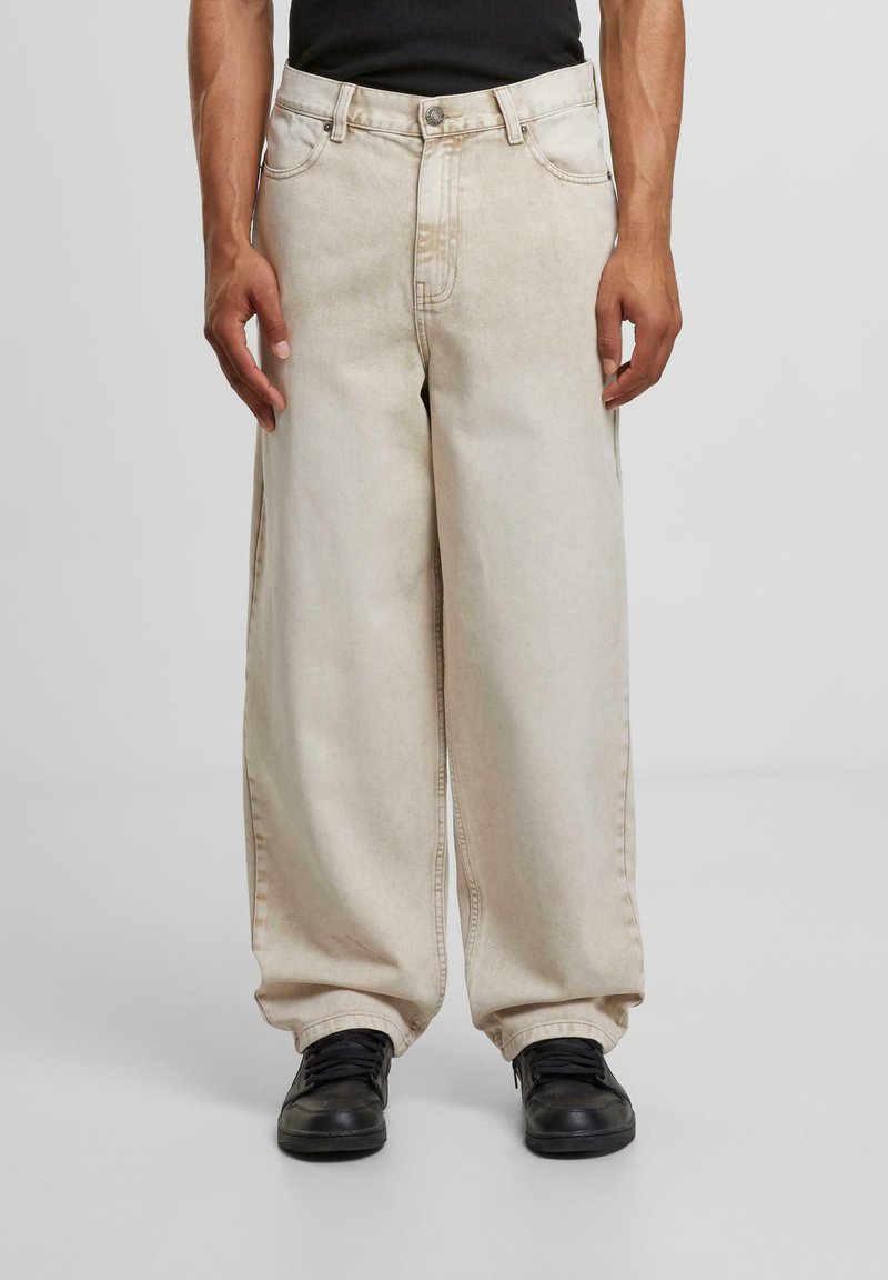 Beige wide-leg jeans made of denim with a faded wash, featuring five pockets and silver hardware. Paired with black sneakers.