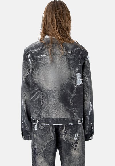 Personne aux longs cheveux portant une veste en jean noire délavée et un pantalon assorti, vue de dos sur un fond gris clair uni.