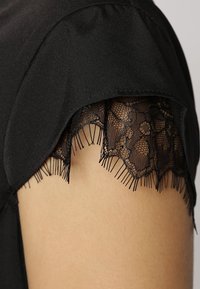 Manche de blouse noire avec un bord en dentelle festonnée, mettant en valeur des motifs floraux et des détails de franges douces. Texture de tissu lisse sur le corps principal.