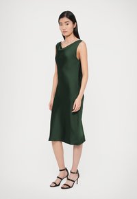 Rochie fără mâneci din satin verde, cu croială lejeră, tiv până la genunchi și un luciu subtil. Asortată cu sandale negre cu barete și toc înalt.