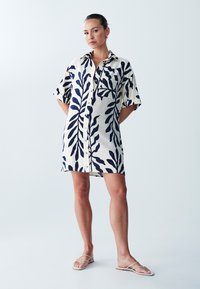 Abito camicia a maniche corte color crema con stampa di foglie blu, dotato di chiusura con bottoni, tasca frontale e vestibilità rilassata.