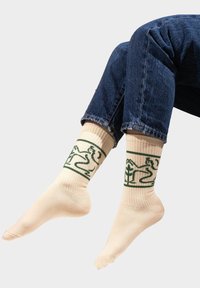 Beżowe skarpetki z zielonymi graficznymi wzorami przedstawiającymi postać i roślinę, z prążkowanym ściągaczem i wzmocnioną piętą w swobodnym środowisku z dżinsami.