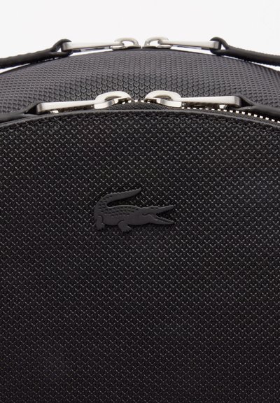 Sac structuré noir avec une finition texturée, des accessoires en argent et un logo Lacoste noir bien visible. Design compact avec une fermeture sécurisée.