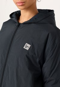 Närbild på en person som bär en svart zip-up-hoodie med ett fyrkantigt märke märkt "LA MAISON AMV" på bröstet.
