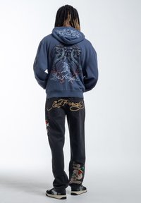 Blauwe hoodie met grafisch ontwerp van slangen en tekst; zwarte jeans met kleurrijke geborduurde accenten en gestikte details; casual schoeisel.