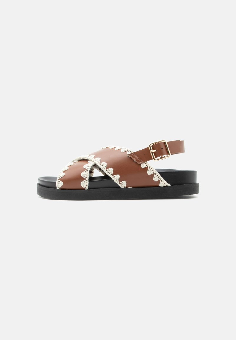 Sandale en cuir marron avec couture en zigzag blanche, semelle noire, brides croisées et sangle de cheville réglable avec boucle dorée.