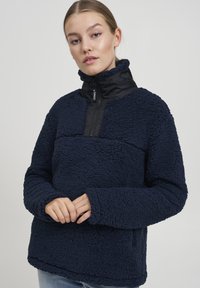 Pullover in pile blu navy con colletto zip nero, superficie testurizzata, maniche lunghe e tasche laterali. Presenta un design moderno e casual.