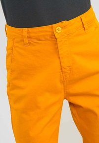 Orange Baumwollhosen mit Knopfverschluss, Gürtelschlaufen und glatter Textur. Der Stoff wirkt leicht und gut verarbeitet.