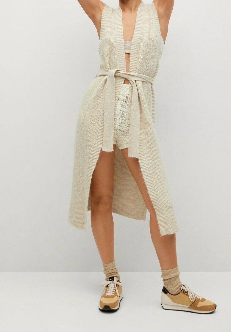 Długi kardigan z przodu otwarty w jasnobeżowym kolorze, wykonany z dzianiny, z paskiem w talii i lekko postrzępionymi krawędziami. Noszony na dopasowanych szortach z dzianiny i trampkach.