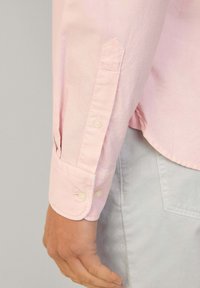 Camicia a maniche lunghe di colore rosa chiaro con polsini appuntiti, dotata di due chiusure a bottone e una texture tessuta liscia. Dettagli delle cuciture visibili sul polsino.