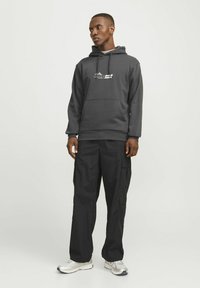 Szary hoodie z przednią kieszenią i logo, zestawiony z czarnymi spodniami cargo z wieloma kieszeniami. Model noszący białe i szare sneakersy.