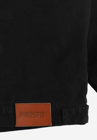 Czarny dżinsowy materiał o teksturowanej powierzchni. Wyróżnia się prostokątną brązową skórzaną metką wytłoczoną napisem "PROSTO". Widoczne detale przeszyć.