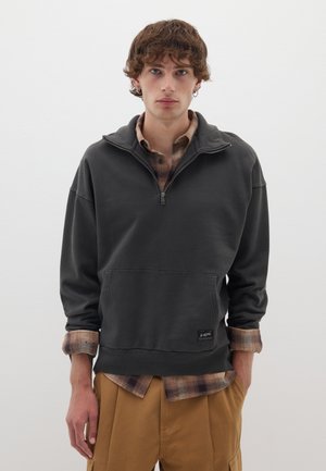 Un jeune homme aux cheveux bouclés porte un sweat-shirt à col zippé gris foncé sur une chemise à carreaux beige et un pantalon beige, se tenant devant un mur blanc uni.